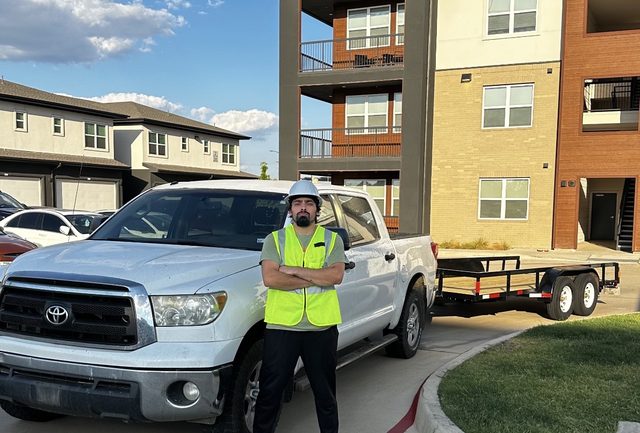 Z&M crew member in a hi-vis vest and hard hat standing beside the white pickup and flatbed trailer outside an apartment building